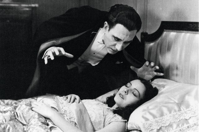Dracula the vampire leans over a woman sleeping in a bed