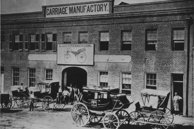 Antique photo of five carriages parked in front of a brick warehouse. Four men stand in the entrance to the building below a sign that reads L. Lichtenberger Carriage Manufactory.