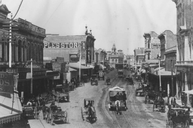 Antique photograph of horse-drawn carriages and biggies, some parked, some driving, along a wide dirt road with brick buildings lining either side.