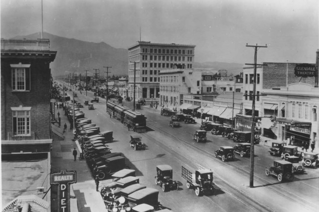 Antique photograph of a wide paved street with an electric trolly car running down the middle. Parked Model T cars and commercial buildings line either side of the street. 