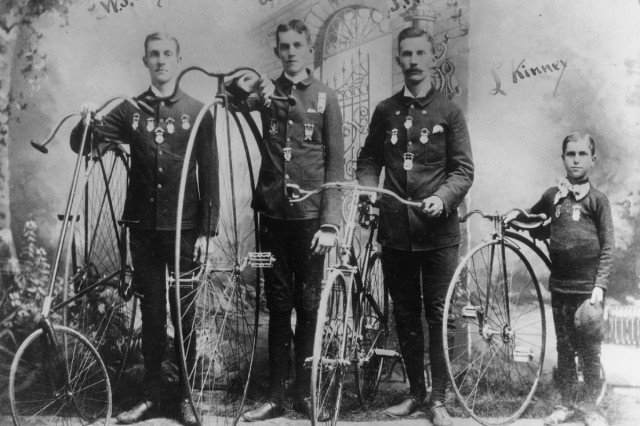 Antique photo of four men with medals pinned to their jackets, standing next to their antique bicycles of varying heights and sizes.