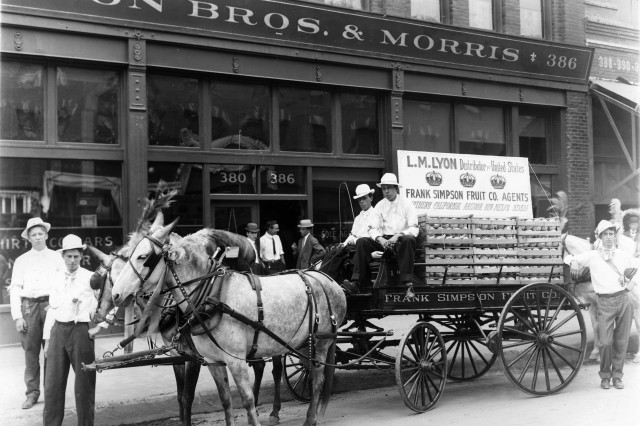 Antique photograph of fruit company wagon drawn by two white horses posing in front of a glass-front store. Crates of fruit fill the wagon bed with a large sign on top that reads Frank Simpson Fruit Co. Agents. Two men stand in front of the horses, two men ride the wagon and one man stands at the back.