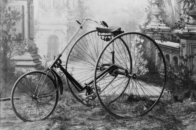 Antique photo staged in an outdoor scene of an antique three-wheeled tricycle with one small wheel in front and two giant wheels in back.