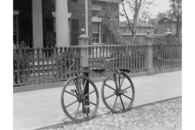 Antique wooden bicycle a cobblestone street, leaning against a post on the sidewalk, with a brick house surrounded by a wooden fence in the background.