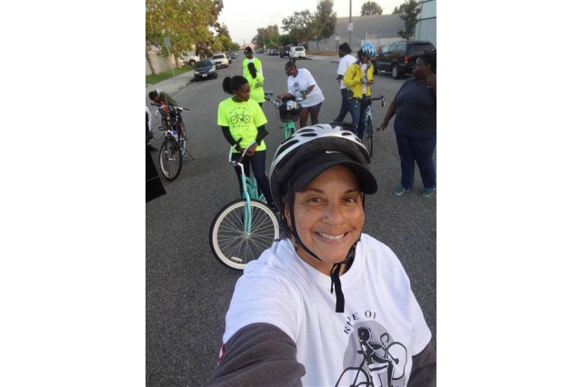 Selfie of a woman of color in a bike helmet, standing in the street in front of a group of bicyclists not paying attention to the camera.