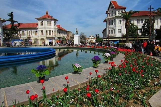 European town square with large, central circular fountain, framed by two white three-story buildings topped with red Spanish tilies