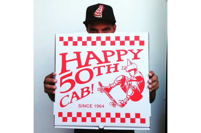 Man in a black baseball cap with a slide of pizza design on it looks over the top of a pizza box (only his eyes are visible) that reads &quot;Happy 50th Cab&quot; in red letters with a design of a skateboarder on the box.