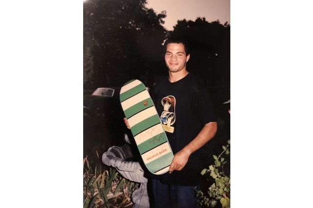 Teenager in a black t-shirt and dark pants, stands in a tree-filled yard, in front of a small, round, cement water fountain, holding a green and white striped skateboard deck.