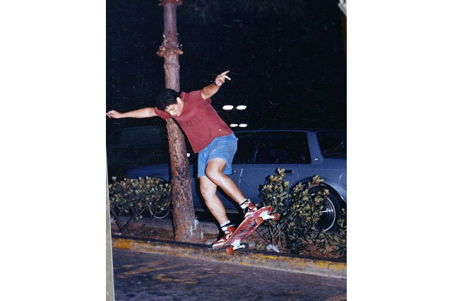 Teenage boy in red t-shirt and jean shorts lifting his skateboard in the air with both feet, his arms outstretched.