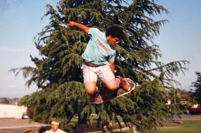 Teenage boy in green t-shirt and white short grabbing his skateboard from behind him, midair with both feet on the board.