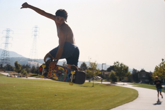 Shirtless skateboarder in shorts, grabbing his skateboard midair from behind, with a grassy park in the background
