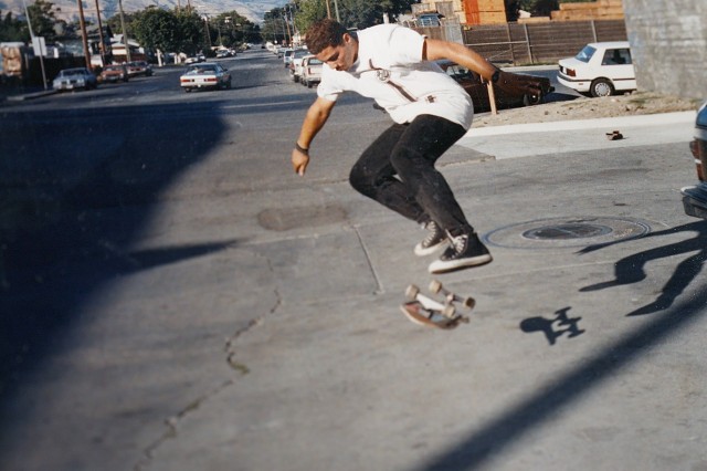 Skateboarder in white t-shirt and black pants, flipping his skateboard in the air, down a wide city street