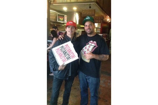 Two men with baseball caps stand side by side with a pizza box