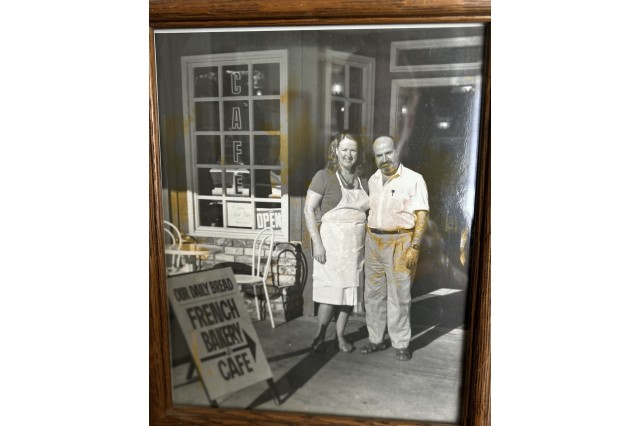 Black and white photo of husband and wife standing in front of their bakery and café beside a sandwich board welcoming in customers