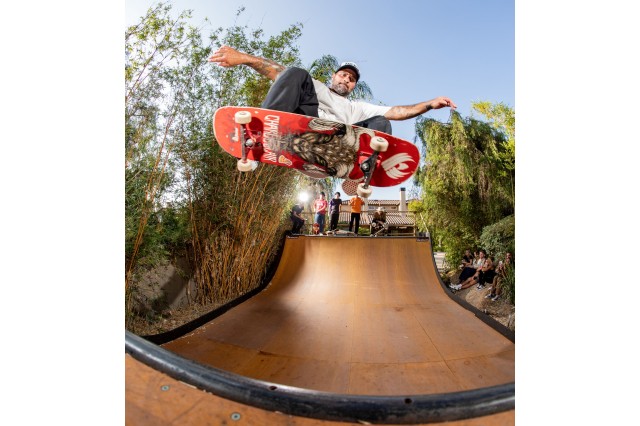 View from below of a bearded man in a baseball cap, white t-shirt and grey pants, skateboarding in midair at the edge of a skate ramp with five other skateboarders watching him from the other side of the ramp.