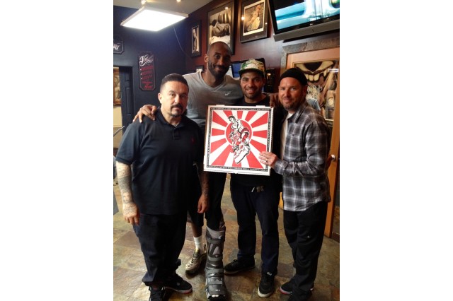 Three men standing around Kobe Bryant, wearing a cast on his left foot. The group is holding up red and white pizza box with a design of a skateboarder on it.