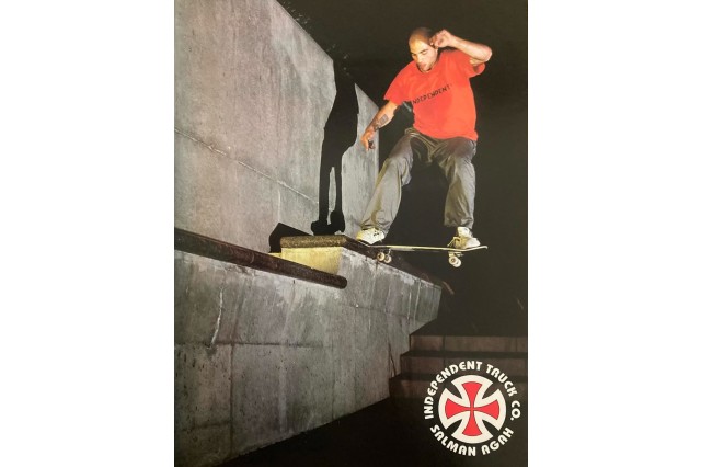 Young man with a shaved head, red t-shirt, black pants, and white sneakers, skateboards at night with the front edge of his board