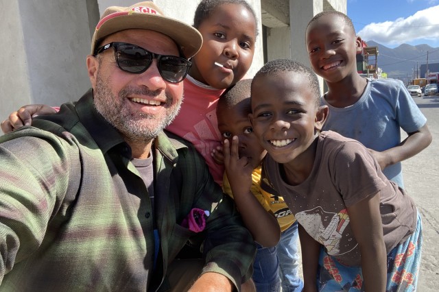 Man with a salt &amp; pepper beard, in a baseball hat, sunglasses and grey plaid shirt, taking a selfie with four young kids crowded around him.