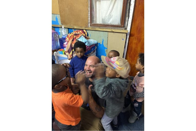 Bearded bald man sitting on the floor inside a small room surrounded by little children. He is giving an enthusiastic double thumbs up to the child in front of him who has tried n his black sunglasses that are too big for his little face.