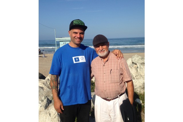 Two men standing on a sidewalk along the beach. The man on the left is tall wearing a baseball hat, blue t-shirt and dark pants, with his arm around a shorter, older man in a baseball hat, red plaid button-down short-sleeved shirt and white pants, holding a black jacket in his left hand. 