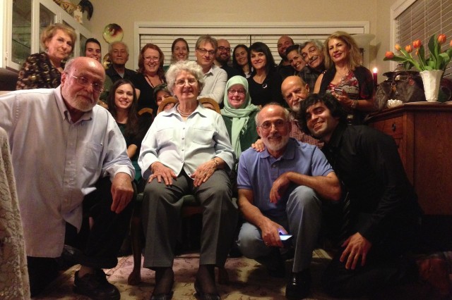 A large, multi-generational family photo in a living room with 13 people standing in back and 7 family members kneeling in front around an older woman sitting in a chair.
