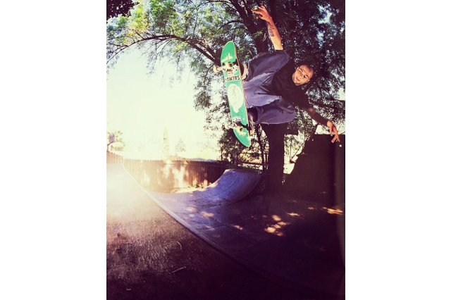 Young man in black t-shirt and pants hangs sideways midair with his arms out and his knees pulled into his chest as he skates high off a ramp on a green skateboard in a small backyard with a tree and fence behind the skate ramp.