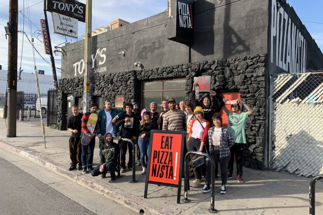 Seventeen young men and women standing behind a red sandwich board sign that says &quot;Eat Pizzanista!&quot;, in front of a black brick restaurant with the Pizzanista! logo painted in large white letters on the side. 