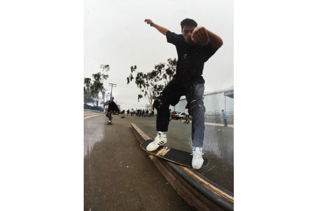 Young male skateboarder skating outside on an overcast day with palm trees in the background, wearing a black t-shirt, jeans, and white sneakers, his arms are up in the air for balance as he looks down at his feet on either side of a black skateboard, straddling a low wooden rail. 