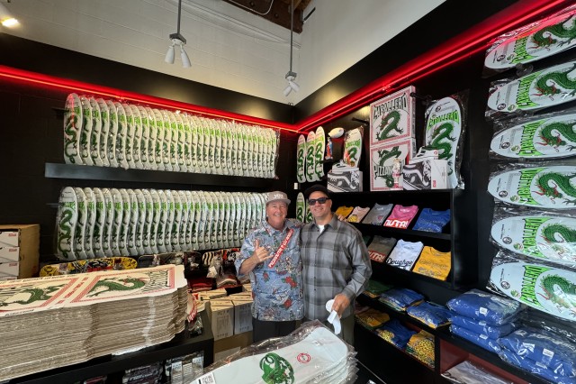 Two men side hugging in a skateboard shop, surrounded by new &quot;Caballero&quot;-branded pizza boxes and skateboard decks, designed with an illustration of a green snake-like dragon. 