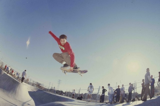 Young man in long-sleeved red shirt and khaki pants with one hand up in teh air and the other crabbing his skateboard as he jumps in midair above a skate ramp.