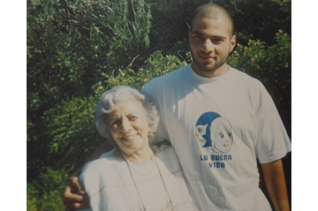 Young man with shaved head, in a white t-shirt with his right arm around his white-haired grandmother who wears a white top and comes up to the man&#039;s shoulder.
