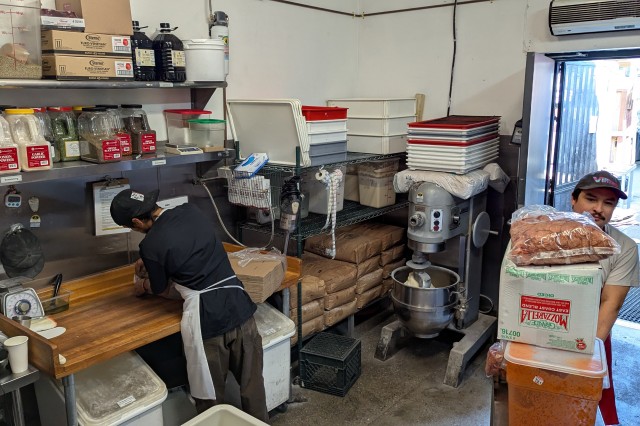Inside a small restaurant kitchen, one man wheels in a metal cart from an open back door with supply boxes, while a second man faces a counter, unpacking cardboard boxes.