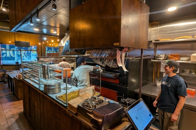 Inside a small restaurant with an open kitchen, two men in white shorts prepping pizzas and sliding them into a large clay oven, while a third man in a black shirt stands off to the side chatting with his busy coworkers.
