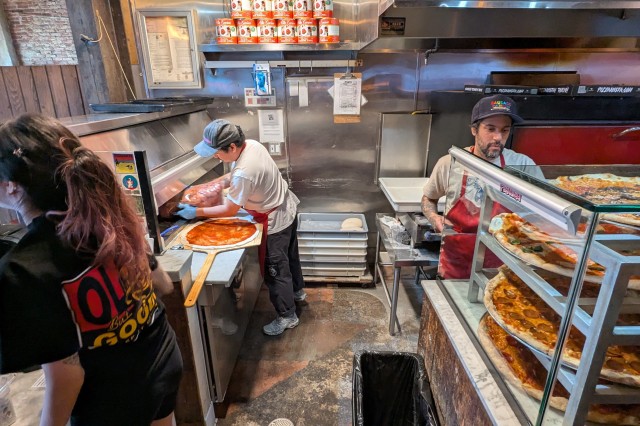 Young woman taking orders at a cash register next to a man placing pepperoni on raw pizza dough, to the right is a man pulling fresh baked pizzas out of an oven and stacking them on cooling racks.