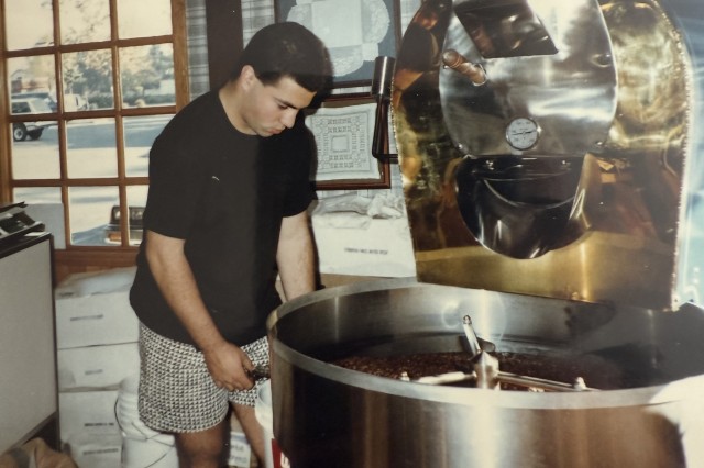 Teenage boy in black t-shirt and black &amp; white shirts turning beans in a large coffee roaster neat a window inside a bakery.