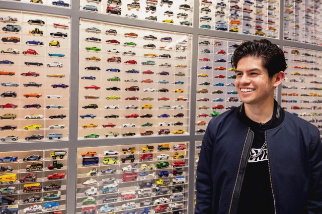A smiling man stands in front of a display of about 800 Hot Wheel and Matchbox cars