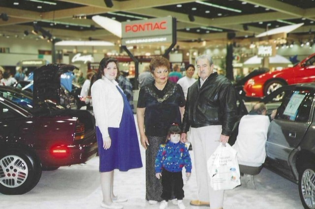 A pregnant woman, an older couple and a young girl stand at a car show.