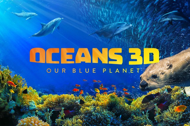 Oceans 3D: Our Blue Planet in yellow-orange capital letters against a background of dolphins swimming in the ocean above a coral reef