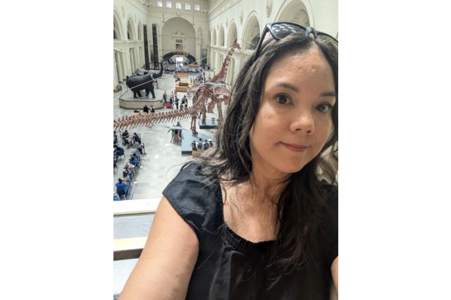 Laura Feucht with black glasses on her head and a black shirt, with a dinosaur skeleton in a large hall behind herith a