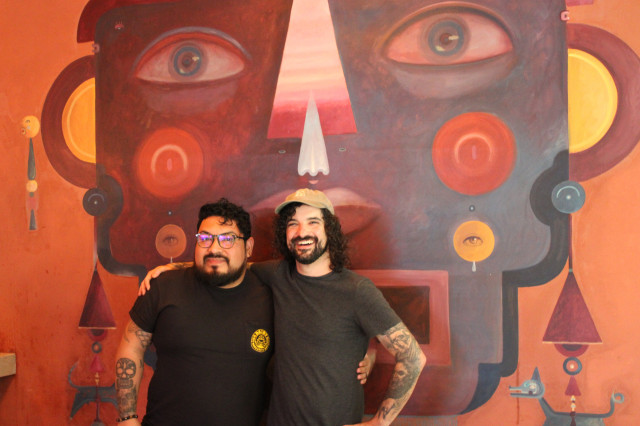 Two men in dark t-shirt standing with an arm around each other's shoulders in front of an abstract art mural of a face.