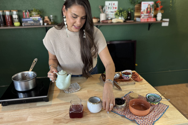 Woman in short-sleeved white top, with long brown hair, leans over a wooden table, holding a white teapot surrounded by bowls full of ingredients.