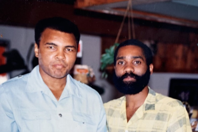 A historical color photograph of a young Percell Keeling with a full black beard standing next to the legendary boxer Muhammad Ali inside the original Simply Wholesome location.