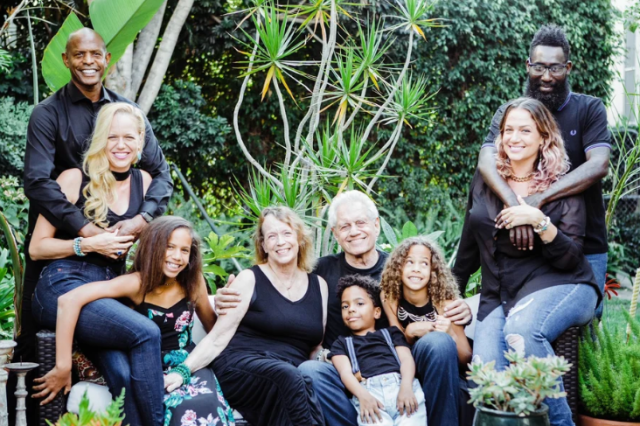 Mixed race, multi-generational family photo taken outdoors in front of lush greenery. Grandparents are seated in the middle with three granddaughters seated below them, and two daughters with their spouses standing on either side.