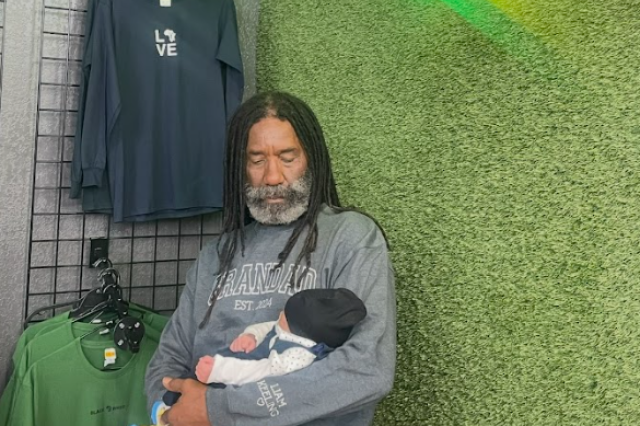 Simply Wholesome founder Percell Keeling sits against a green textured wall, tenderly cradling his infant grandson, Liam. Percell is wearing a grey sweatshirt that reads "GRANDAD EST. 2024" on the chest, with "LIAM KEELING" embroidered on the sleeve.