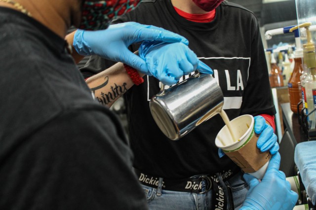 Two baristas dressed in black, pouring steamed milk into a paper cup.
