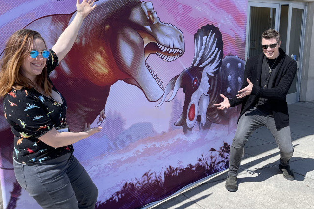 Annie with arms raised towards a mural of two dinosaurs facing each other, with Ted Rechlin on the side lunging with open hands towards the mural