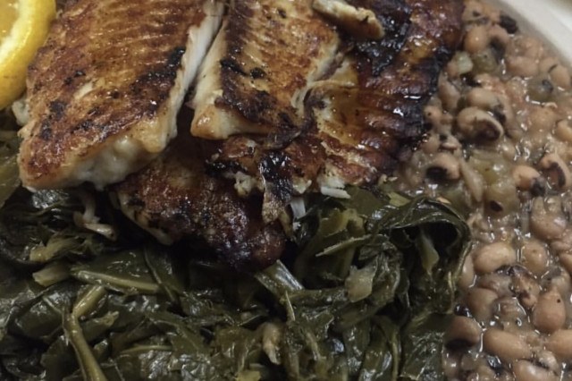 A close-up shot of two blackened tilapia fillets with a dark, flavorful crust, served over a bed of collard greens with a side of black-eyed peas and orange garnishes.