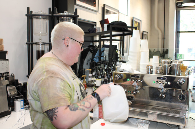 A Blonde haired barista pours  milk into a cup at the counter of the Kavat coffee shop.