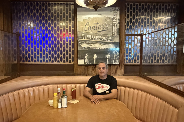 Man sits in a large retro style restaurant booth below a photo of the original Canter's Deli.