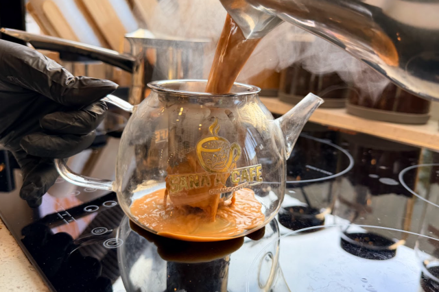 Close-up of a gloved hand holding a glass teapot while a hot, creamy brown liquid is poured into it from a metal saucepan, with steam rising and jars of ingredients visible in the background.